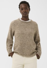 Femme portant un pull en tricot beige sur une chemise blanche et un pantalon crème, se tenant avec les mains dans les poches devant un fond blanc uni.
