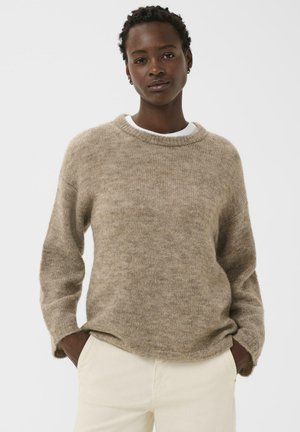 Woman wearing a beige knit sweater over a white shirt and cream pants, standing with hands in pockets against a plain white background.