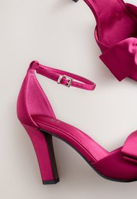 Vaaleanpunaiset satiinikorolliset kengät, joissa on nilkkaremmi ja rusetti etuosassa. Tyylikäs muotoilu, kiiltävä pinta ja kapea korko.