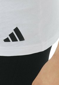 Zbliżenie na osobę w białej bluzce z czarnym logo w trzy paski i czarnych spodniach, pokazującą bok torsu i ramienia.