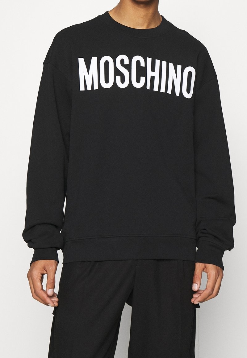 Sweat-shirt noir en tissu doux, arborant un grand logo blanc "MOSCHINO" sur la poitrine. Coupe légèrement surdimensionnée avec des poignets côtelés.