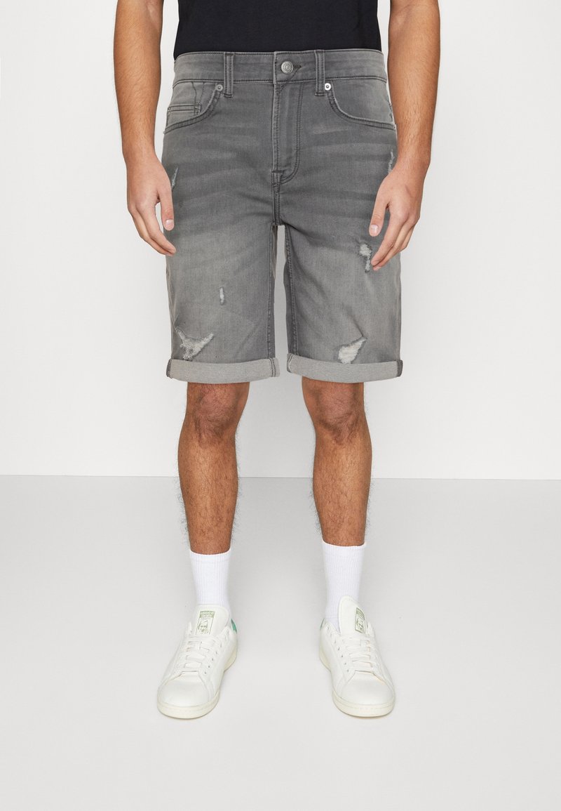 Shorts en jean gris avec des ourlets retroussés, présentant des détails usés et un design classique à cinq poches, associés à des baskets blanches et des chaussettes.