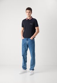 Ναυτικό πόλο μπλουζάκι κοντό μανίκι με κόκκινο και άσπρο ριγέ γιακά; σε συνδυασμό με ανοιχτό γαλάζιο τζιν και άσπρα αθλητικά παπούτσια σε φωτεινό φόντο.