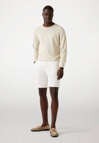 Lys beige langærmet sweater og hvide shorts, parret med brune ruskinds mokasiner. Simpelt design, letvægtstof og minimal detaljer.