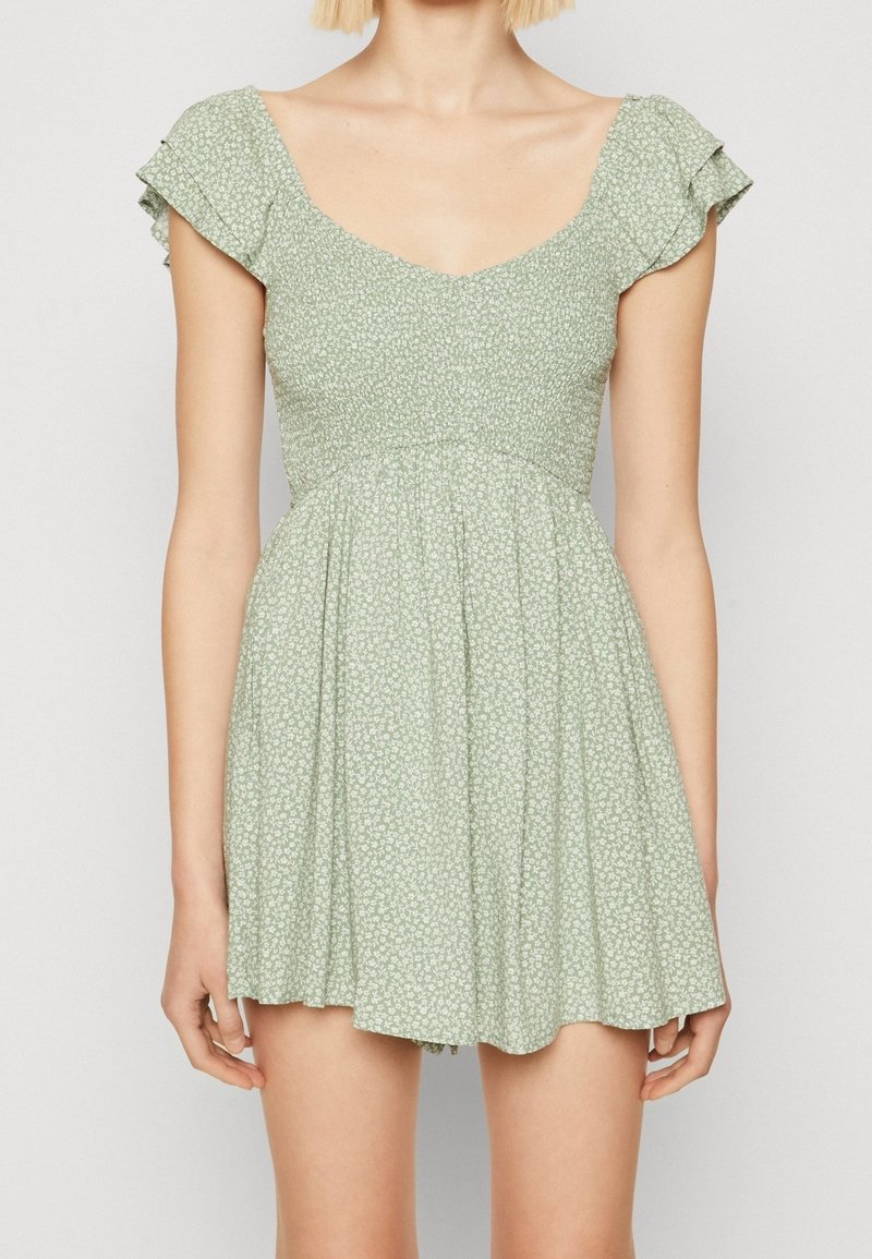 Femme portant une robe courte à fleurs vert clair avec une taille froncée et des manches courtes volantées, se tenant contre un fond uni.