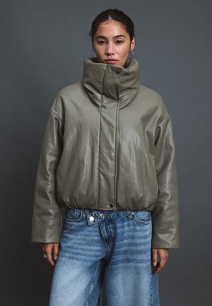 Jeune femme portant une veste matelassée vert olive avec un col montant et un jean bleu, debout devant un fond gris foncé.