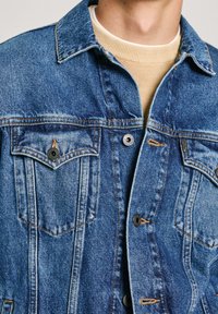 Chaqueta de denim en un azul medio con dos bolsillos en el pecho, botones negros y costuras en contraste. Presenta un cuello clásico y una tela texturizada.