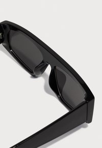 Schwarze Sonnenbrille mit schlankem Design, rechteckiger Form und dunklen Gläsern. Die Fassung hat ein glänzendes Finish und ausgeprägte, kantige Kanten.