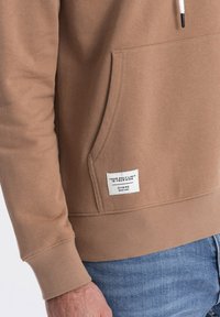 Felpa marrone con tasca a marsupio, polsini e orlo a coste, con etichetta bianca con testo, realizzata in tessuto morbido.