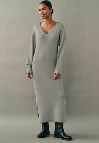 Robe en tricot grise longue avec un décolleté en V profond, texture côtelée et fentes sur les côtés, associée à des bottes noires montantes.