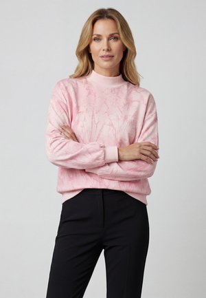 Donna con capelli biondi che indossa un maglione rosa chiaro a maniche lunghe con motivo e pantaloni neri, in piedi con le braccia incrociate contro uno sfondo grigio.