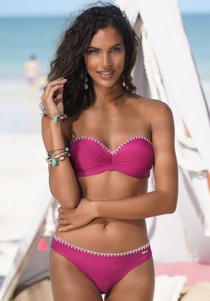 Magenta bikinový set s bandeau topem a bílým zigzagovým lemováním a odpovídajícími spodními díly. Modelka doplnila vzhled o vrstvené náramky a náušnice.