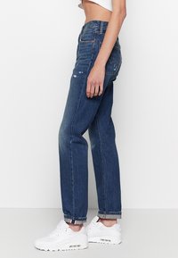 Femme portant un jean bleu taille haute, coupe droite, avec des revers roulés et des baskets blanches, debout de profil sur un fond blanc uni.