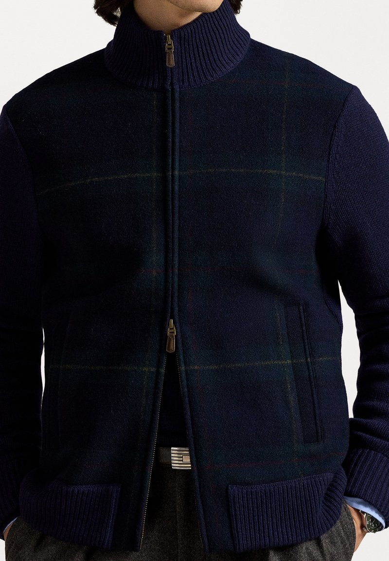 Navy zip-up vest with high collar, featuring a plaid design, ribbed knit accents, and front pockets. Made from a soft wool blend.