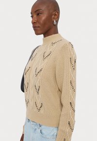 Beezist kampsun kõrge kaelusega, millel on kabelkootud disain ja kontrastsed mustad ja valged dekoratiivsed aktsendid külgedel.