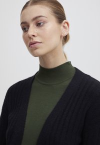 Svart ribbad cardigan över en grön polotröja. Cardiganen har en djup V-ringning och en mjuk textur, som kontrasterar med den släta polotröjan.