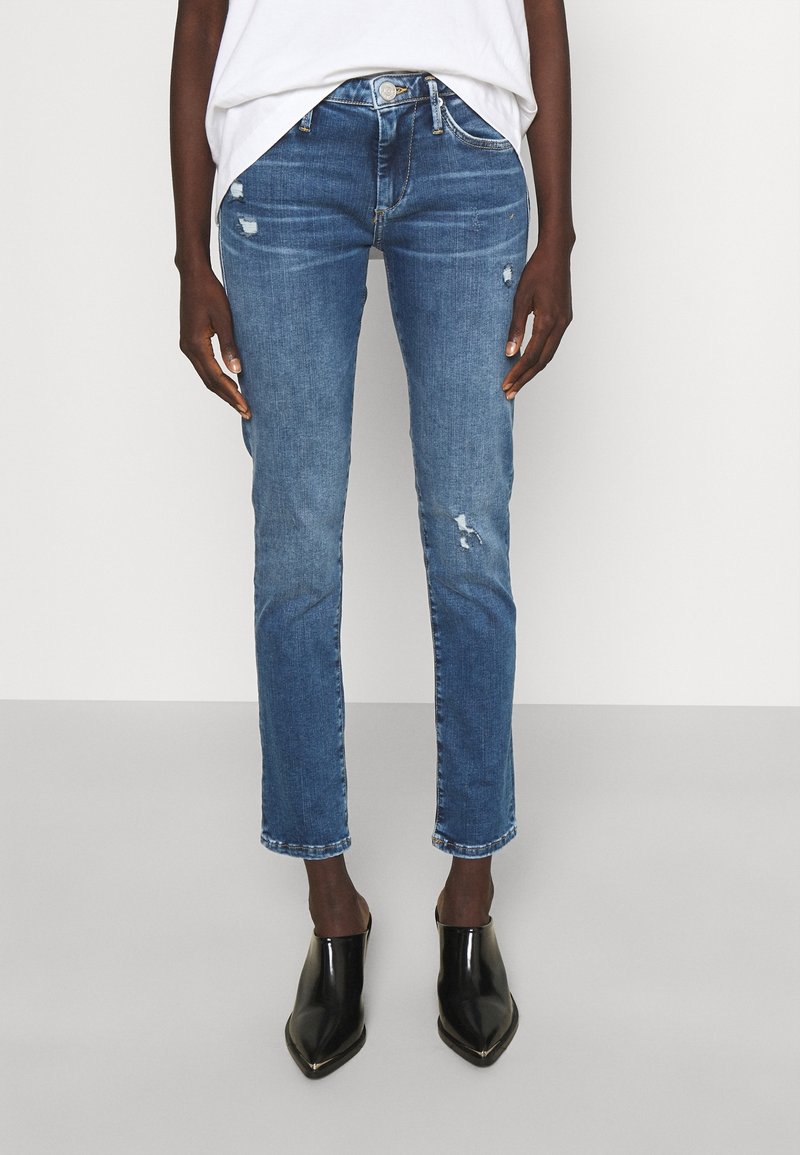 Jeans azul ajustados con ligeros desgastes, combinados con una camiseta blanca y botines negros de punta afilada y brillo, en una persona de pie.