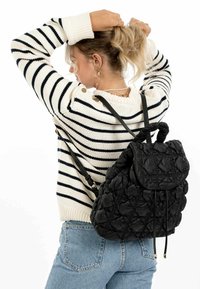 Sac à dos noir matelassé avec un rabat arrondi, fermeture à cordon et poignée tressée sur le dessus. Porté sur le dos, associé à un pull rayé.