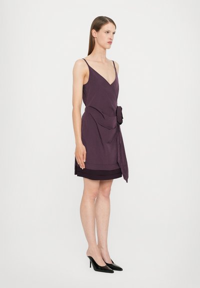 Purple sleeveless dress featuring a V-neck, a tie detail at the waist, and layered hem. Worn with black pointed-toe heels.