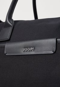 Čierna textilná taška s koženými akcentmi, štruktúrovaný tvar a strieborné logo "JOOP!" na obdĺžnikovom štítku. Celkovo hladká textúra.