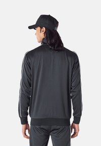 Giacca sportiva nera con colletto alto e strisce bianche sui lati. Realizzata in tessuto liscio e leggero, presenta un orlo aderente e pantaloni abbinati.