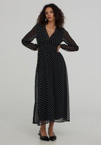Czarna maxi sukienka w kropki z długimi przezroczystymi rękawami, dekoltem w kształcie litery V, dopasowaną talią i zwiewnym dołem, uzupełniona delikatnymi czarnymi szpilkami.