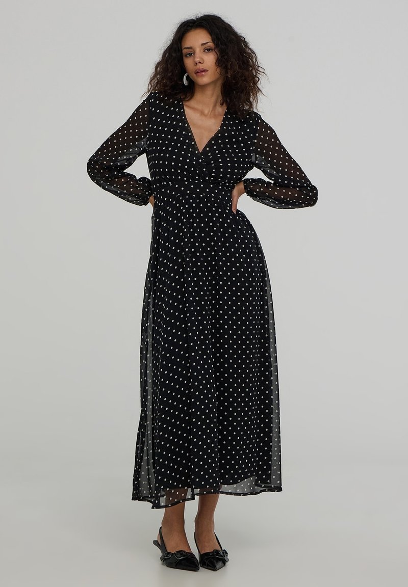 Czarna maxi sukienka w kropki z długimi przezroczystymi rękawami, dekoltem w kształcie litery V, dopasowaną talią i zwiewnym dołem, uzupełniona delikatnymi czarnymi szpilkami.