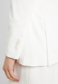 La manica del blazer bianco presenta un tessuto liscio, un polsino a bottone singolo con quattro bottoni coordinati e una silhouette sartoriale.