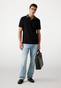 Black polo shirt with patterned collar, paired with light blue jeans and black shoes. Model holds a black leather bag.