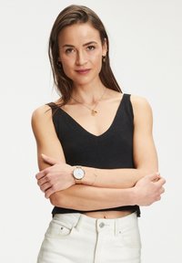 Schwarzes geripptes Tanktop mit V-Ausschnitt, kombiniert mit weißen Jeans. Die Accessoires umfassen eine goldene Uhr und gestapelte goldene Schmuckstücke. Sauberer Hintergrund.