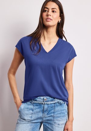 Frau mit langen dunklen Haaren, die silberne Creolen, ein blaues V-Ausschnitt-Kurzarmshirt und hellblaue, gemusterte Jeans trägt, die Hände in den Taschen.