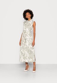 Abito senza maniche, lungo fino alla caviglia, in tessuto leggero con una stampa floreale tropicale verde, caratterizzato da una vita legata e spacco laterale, abbinato a tacchi beige.