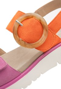 Bunt gefärbte Sandale mit rosa, orange und beige Wildlederriemen, ausgestattet mit einem runden, durchsichtigen Akzent und einer weißen, strukturierten Plattformsohle.
