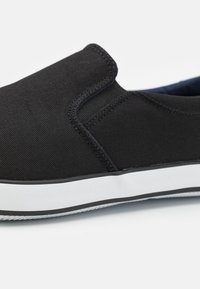Black canvas slip-on shoe featuring a textured upper, elastic side panels, and a white rubber sole with a black stripe.