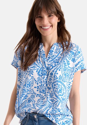 Smilende kvinde med langt brunt hår iført en hvid kortærmet bluse med et blåt paisley-mønster og blå jeans.