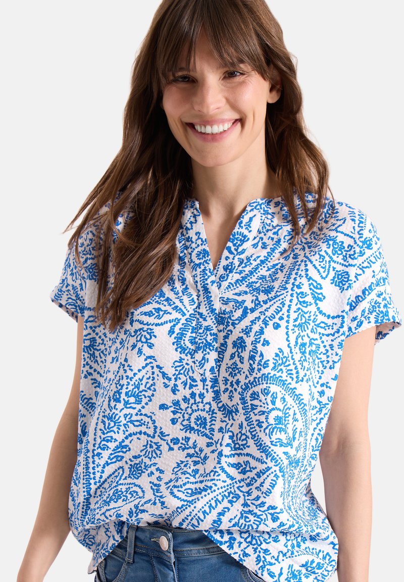 Femme souriante aux longs cheveux bruns portant un chemisier blanc à manches courtes avec un motif paisley bleu et un jean bleu.