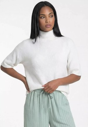 Kobieta o długich czarnych włosach, ubrana w biały sweter z krótkim rękawem i golfem oraz miętowo-zielone spodnie w prążki ze sznurkiem.