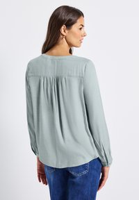 Blouse vert clair à manches longues avec un col rond, détails de plis au dos et manches à boutons, associée à un jean bleu.