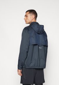 Veste coupe-vent marine avec capuche, présentant un design de dos superposé, des poignets élastiques et un logo réfléchissant dans le bas du dos.