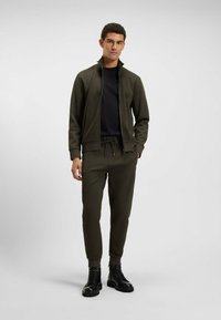 Olivgrüne Zip-Jacke mit hohem Kragen, kombiniert mit passenden schmal zulaufenden Jogginghosen und schwarzen Schnürstiefeln. Glattes Material und minimale Details.