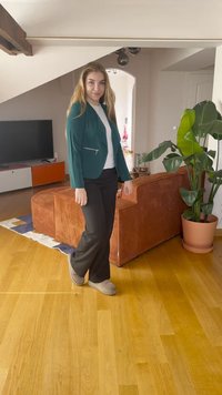 Zeleni blejzer z zadrgo, čez belo srajco, črne hlače in bež copate. Ozadje vključuje teksturirano oranžno kavč in sobne rastline.