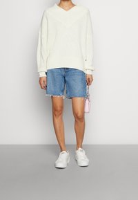 Weißer, gestrickter Pullover mit V-Ausschnitt und gerippten Bündchen, kombiniert mit blauen Denim-Shorts mit fransigen Säumen und weißen Sneakers. Kleine rosa Handtasche.