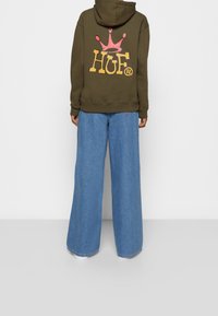 Felpa verde con cappuccio e un grande logo colorato "HUF" sulla schiena, abbinata a jeans in denim azzurro a gamba larga.