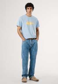 Šviesiai mėlynas medvilninis marškinėliai su geltonu "Pepe Jeans London" logotipu, derinami su šviesiai mėlynomis džinsinėmis kelnėmis ir smėlio spalvos slip-on batais.