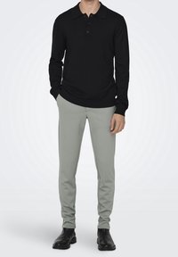 Polo shirt nero a maniche lunghe con colletto e tre bottoni, abbinato a pantaloni slim-fit grigio chiaro e stivali alla caviglia neri.