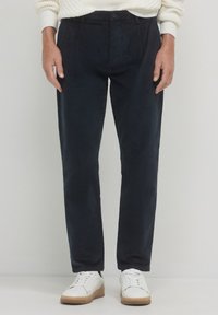 Pantaloni in velluto a coste blu navy con taglio dritto, caratterizzati da pieghe frontali e chiusura a bottone. Abbinati a sneakers bianche per un look casual.