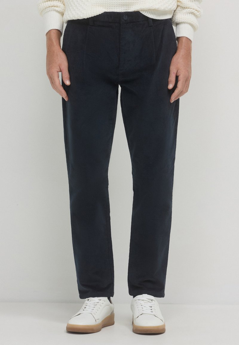 Pantaloni in velluto a coste blu navy con taglio dritto, caratterizzati da pieghe frontali e chiusura a bottone. Abbinati a sneakers bianche per un look casual.