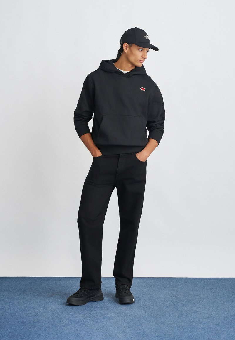 Homme portant un sweat à capuche noir avec un petit logo rouge, un pantalon noir, des baskets noires et une casquette noire, se tenant sur un tapis bleu contre un mur blanc.