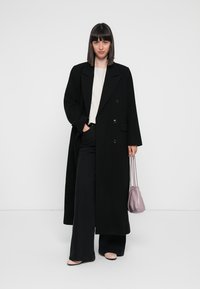 Lungo cappotto nero con vestibilità oversize, design doppiopetto e ampi rever. Abbinato a pantaloni scuri e a una maglietta chiara. Tiene in mano una borsa lilla.