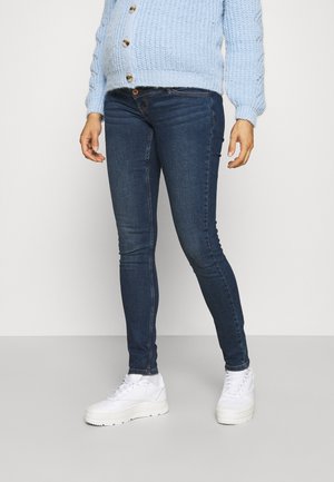 Persoon die donkere skinny jeans, witte sneakers en een lichtblauwe grofgebreide, met knopen afgewerkte trui draagt en staat op een effen achtergrond.